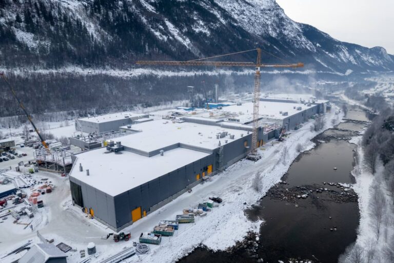 Luftfoto av en stor industriell bygningskonstruksjon i et snødekt landskap. Bygningen ligger ved en elv, omgitt av fjell og skog. Flere byggekraner og maskiner er synlige på området.